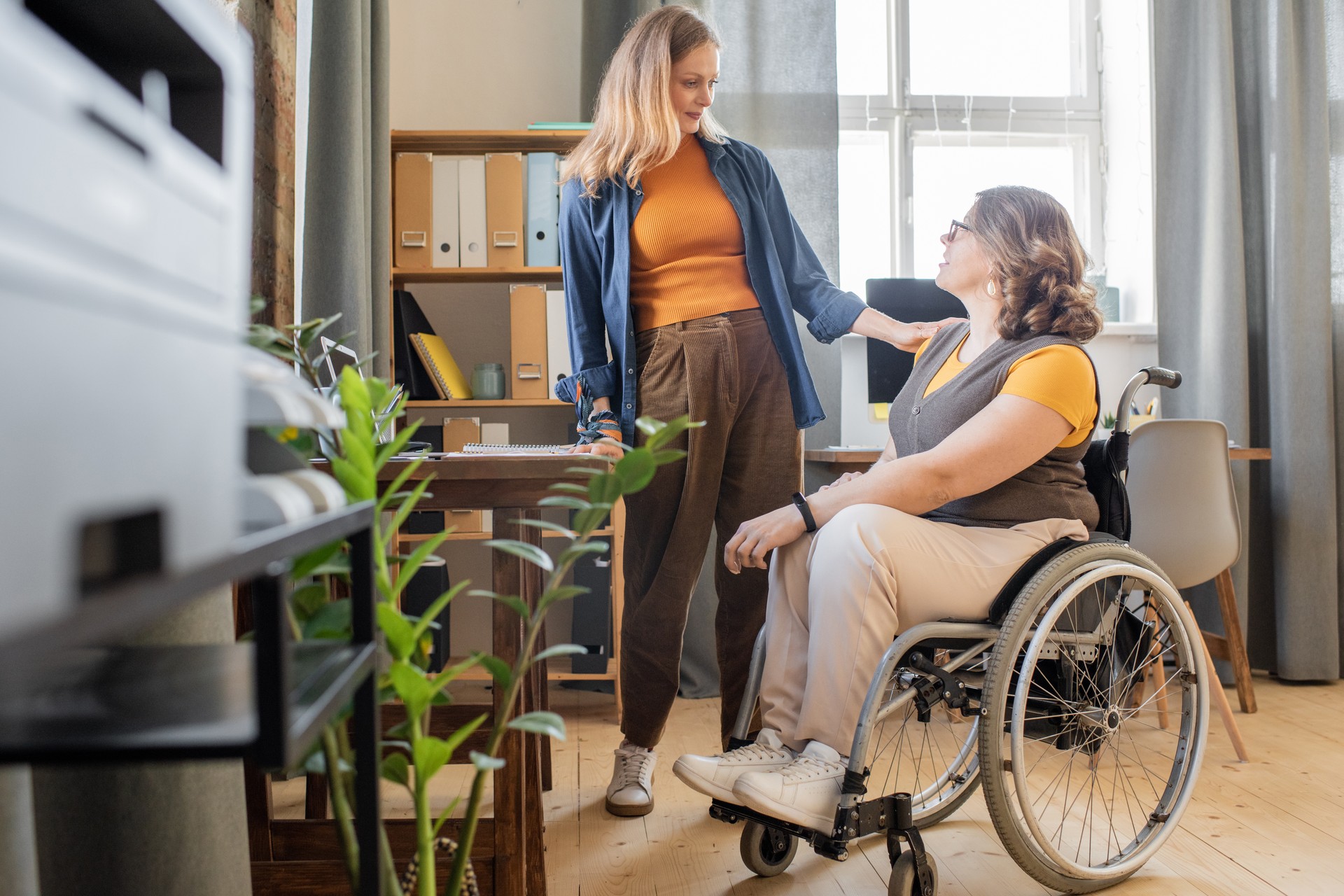 Young black woman in casualwear looking at her disable friend in wheelchair