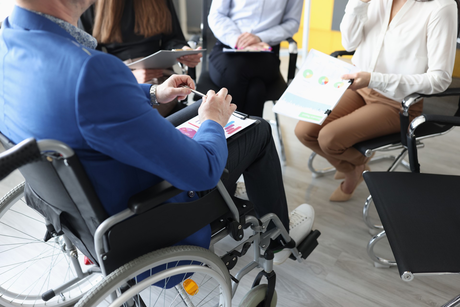 Disabled man with documents in hands sitting in wheelchair against background of colleagues closeup